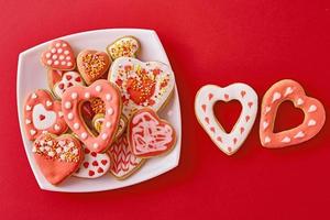 Decorated heart shape cookies in white plate and two cookies on the red background, top view. Valentines Day food concept photo