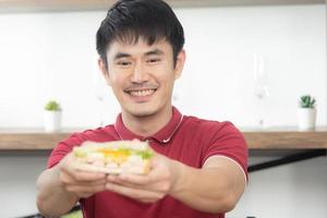 The business smiling man with casual  red t-shirt having breakfast and eating sandwich, Young man cooking food and drink in the loft style kitchen room photo