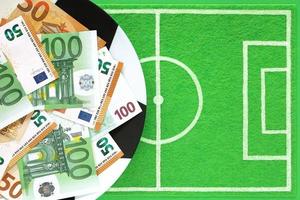 Euro banknotes 50 and 100 on plate painted like soccer ball, dish mat background like small football field made of green felt. Sports betting, soccer bets, football match results, gambling, money. photo