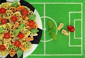 Tricolor pasta in form of football boots, balls and cups on plate like soccer ball, dish mat like football field. Italy flag colors in green basil, white pasta and red cherry tomato. Goal concept. photo