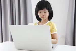 una pequeña estudiante asiática estudia en línea usando una computadora portátil en casa foto