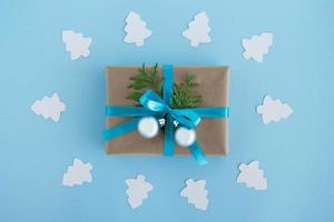 Gift box wrapped of craft paper, blue ribbon and decorated fir branches and silver Christmas balls with white paper fir tree around on the blue background, top view. Christmas present. photo