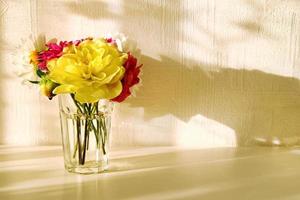 Glass vase with yellow, white, pink and red peonies on the white table in bright sunny light. photo