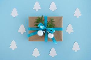 Gift box wrapped of craft paper, blue ribbon and decorated fir branches and silver Christmas balls with white paper fir tree around on the blue background, top view. Christmas present. photo