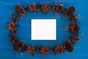 Top view on frame from Christmas lights, pine cones and white sheet of paper on the blue wooden background with copy space. photo