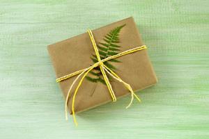Gift box wrapped of craft paper and white and yellow ribbon with fern leaf on a green wooden background, top view. photo