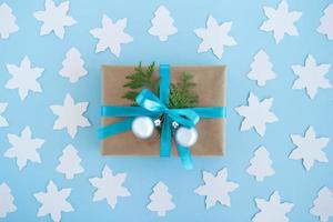 Gift box wrapped of craft paper, blue ribbon and decorated fir branches and silver Christmas balls on the blue background with white paper fir tree and star, top view. Christmas present. photo