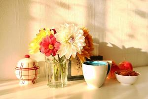 Still life with glass vase with colorful flowers of peonies, cup of tea, apple jam, apples and sugar bowl on the white table in bright sunny light. photo