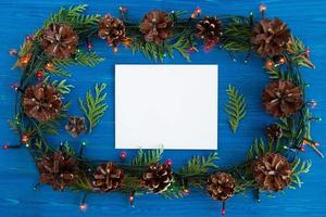 Top view on frame from Christmas lights, fir branches, pine cones and white sheet of paper on the blue wooden background with copy space. photo