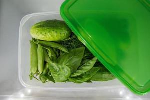 Green vegetable - basil, dill, sorrel, peas, cucumber - in the plastic food container on a shelf of a fridge. photo
