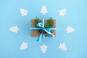 Gift box wrapped of craft paper, blue and white ribbons and decorated fir branches and pinecones on the blue background with white paper fir tree, top view. Christmas present. photo