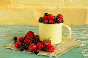 una taza de metal con fresas, frambuesas y grosellas negras en las servilletas de tela de saco sobre la mesa de madera azul-blanca sobre el fondo de la pared de ladrillo. foto