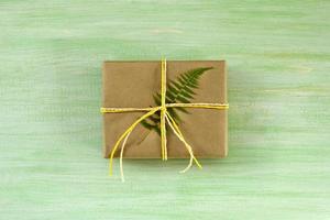 caja de regalo envuelta en papel artesanal y cinta blanca y amarilla con hoja de helecho sobre un fondo de madera verde, vista superior. foto