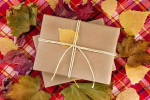 Top view on a gift boxes wrapped of craft paper and white ribbons with dry colorful leaves, on a red checkered towel. Autumn still life. photo