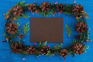 Top view on frame from Christmas lights, fir branches, pine cones and brown sheet of paper on the blue wooden background with copy space. photo