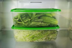 Two plastic food containers with green vegetable - salad, basil, dill, sorrel, peas, cucumber, pepper - on a shelf of a fridge photo