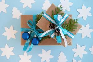 dos cajas de regalo envueltas en papel artesanal, cinta azul y blanca y ramas de abeto decoradas, bolas de navidad azules y piñas en el fondo azul con abeto de papel y estrella, vista superior. foto