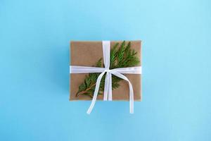 Gift box wrapped of craft paper, white ribbon and decorated fir branch on the blue background, top view. Christmas present. photo