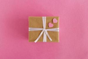 Gift box wrapped of craft paper and white ribbon with two pink hearts on the pink background, top view. photo