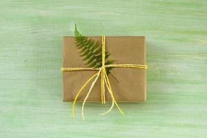 Gift box wrapped of craft paper and white and yellow ribbon with fern leaf on a green wooden background, top view. photo