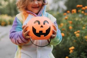 cute little girl in a rainbow unicorn costume for Halloween goes to collect sweets in a pumpkin basket in a residential area. child walks in the outdoor. trick or treat. lifestyle. kigurumi photo