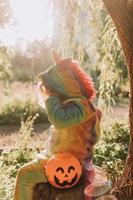 little girl in a rainbow unicorn Halloween costume and a dachshund in a dress with a pumpkin basket for sweets are sitting on stump at forest sunset. fabulous wonderful magical forest. space for text photo