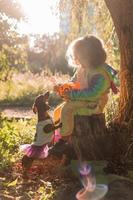 little girl in a rainbow unicorn Halloween costume and a dachshund in a dress with a pumpkin basket for sweets are sitting on stump at forest sunset. fabulous wonderful magical forest. space for text photo