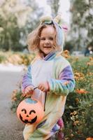 linda niñita con un disfraz de unicornio arcoiris para halloween va a recoger dulces en una canasta de calabaza en una zona residencial. el niño camina al aire libre. truco o trato. estilo de vida. kigumi foto