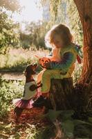 little girl in a rainbow unicorn Halloween costume and a dachshund in a dress with a pumpkin basket for sweets are sitting on stump at forest sunset. fabulous wonderful magical forest. space for text photo