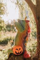 little girl in a rainbow unicorn Halloween costume and a dachshund in a dress with a pumpkin basket for sweets are sitting on stump at forest sunset. fabulous wonderful magical forest. space for text photo