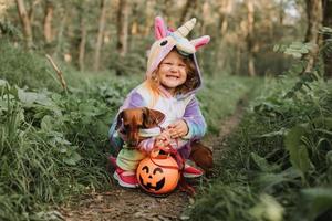 little girl in a rainbow unicorn Halloween costume and a dachshund in a dress with a pumpkin basket for sweets are sitting on stump at forest sunset. fabulous wonderful magical forest. space for text photo