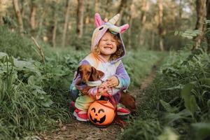 una niña con un disfraz de halloween de unicornio arcoíris y un dachshund con un vestido con una canasta de calabaza para dulces están sentados en un tocón al atardecer del bosque. fabuloso maravilloso bosque mágico. espacio para texto foto