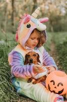 little girl in a rainbow unicorn Halloween costume and a dachshund in a dress with a pumpkin basket for sweets are sitting on stump at forest sunset. fabulous wonderful magical forest. space for text photo