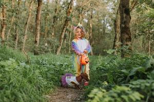 little girl in a rainbow unicorn Halloween costume and a dachshund in a dress with a pumpkin basket for sweets are sitting on stump at forest sunset. fabulous wonderful magical forest. space for text photo