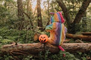 little girl in a rainbow unicorn Halloween costume with a pumpkin basket for sweets is sitting on a stump at a forest sunset. a fabulous wonderful magical forest. space for text. High quality photo