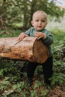 portrait of cute baby boy holding on to a log against the background of a green forest. pumpkin basket for sweets in the foreground. walking and playing outdoor. Halloween concept. High quality photo