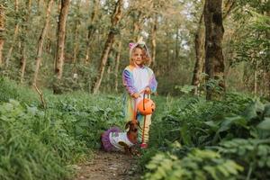 little girl in a rainbow unicorn Halloween costume and a dachshund in a dress with a pumpkin basket for sweets are sitting on stump at forest sunset. fabulous wonderful magical forest. space for text photo