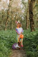 little girl in a rainbow unicorn Halloween costume and a dachshund in a dress with a pumpkin basket for sweets are sitting on stump at forest sunset. fabulous wonderful magical forest. space for text photo