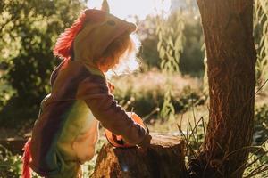 una niña con un disfraz de halloween de unicornio arcoíris y un dachshund con un vestido con una canasta de calabaza para dulces están sentados en un tocón al atardecer del bosque. fabuloso maravilloso bosque mágico. espacio para texto foto