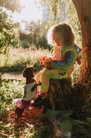 little girl in a rainbow unicorn Halloween costume and a dachshund in a dress with a pumpkin basket for sweets are sitting on stump at forest sunset. fabulous wonderful magical forest. space for text photo