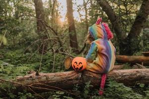 little girl in a rainbow unicorn Halloween costume with a pumpkin basket for sweets is sitting on a stump at a forest sunset. a fabulous wonderful magical forest. space for text. High quality photo