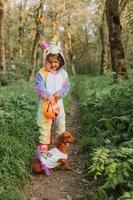 little girl in a rainbow unicorn Halloween costume and a dachshund in a dress with a pumpkin basket for sweets are sitting on stump at forest sunset. fabulous wonderful magical forest. space for text photo