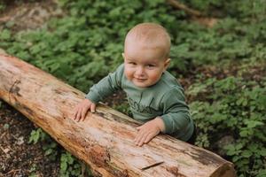 portrait of cute baby boy holding on to a log against the background of a green forest. pumpkin basket for sweets in the foreground. walking and playing outdoor. Halloween concept. High quality photo