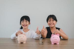 dos niñas asiáticas divirtiéndose para poner monedas en una alcancía juntas, niños ahorrando dinero para el concepto futuro foto