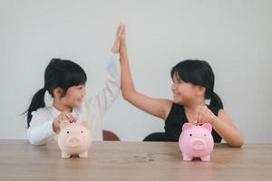 dos niñas asiáticas divirtiéndose para poner monedas en una alcancía juntas, niños ahorrando dinero para el concepto futuro foto