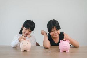 dos niñas asiáticas divirtiéndose para poner monedas en una alcancía juntas, niños ahorrando dinero para el concepto futuro foto