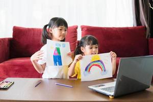Two asian child girl students study online with teacher by video call together. Siblings are homeschooling with computer laptop during quarantine due to Covid 19 pandemic. photo