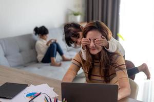 woman sits by the table at home office during lockdown, working on laptop. Playful child distracts from work, covering her mother's eyes, kid making noise and asking attention from busy mom photo