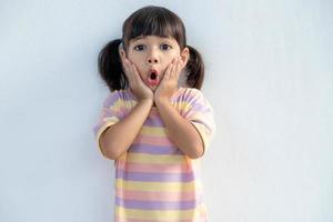 retrato de una niña pequeña sorprendida y linda que se encuentra aislada sobre fondo blanco. mirando a la cámara. manos cerca de la boca abierta foto