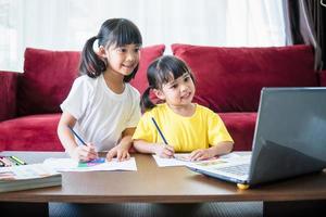 Two asian child girl students study online with teacher by video call together. Siblings are homeschooling with computer laptop during quarantine due to Covid 19 pandemic. photo